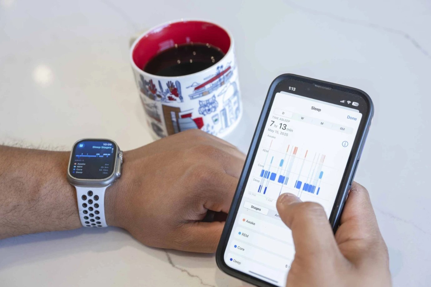 A person’s hands resting on a table, with an Apple Watch on the left wrist and right hand holding an iPhone displaying sleep date. A University of Arizona mug of coffee sits on the table